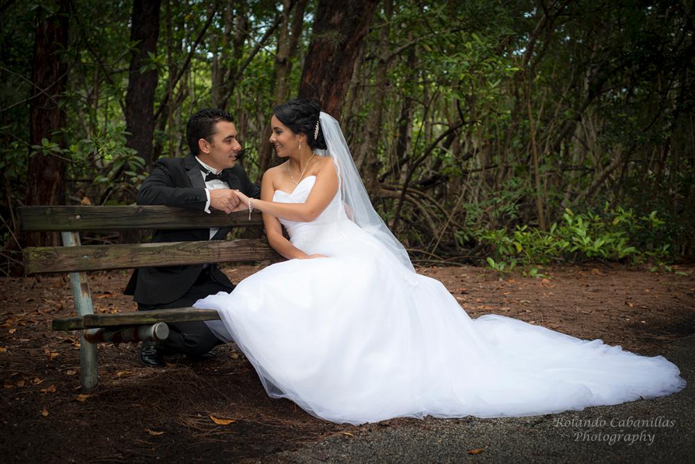 Bride in a white gown and groom in a tuxedo holding hands on a park bench
